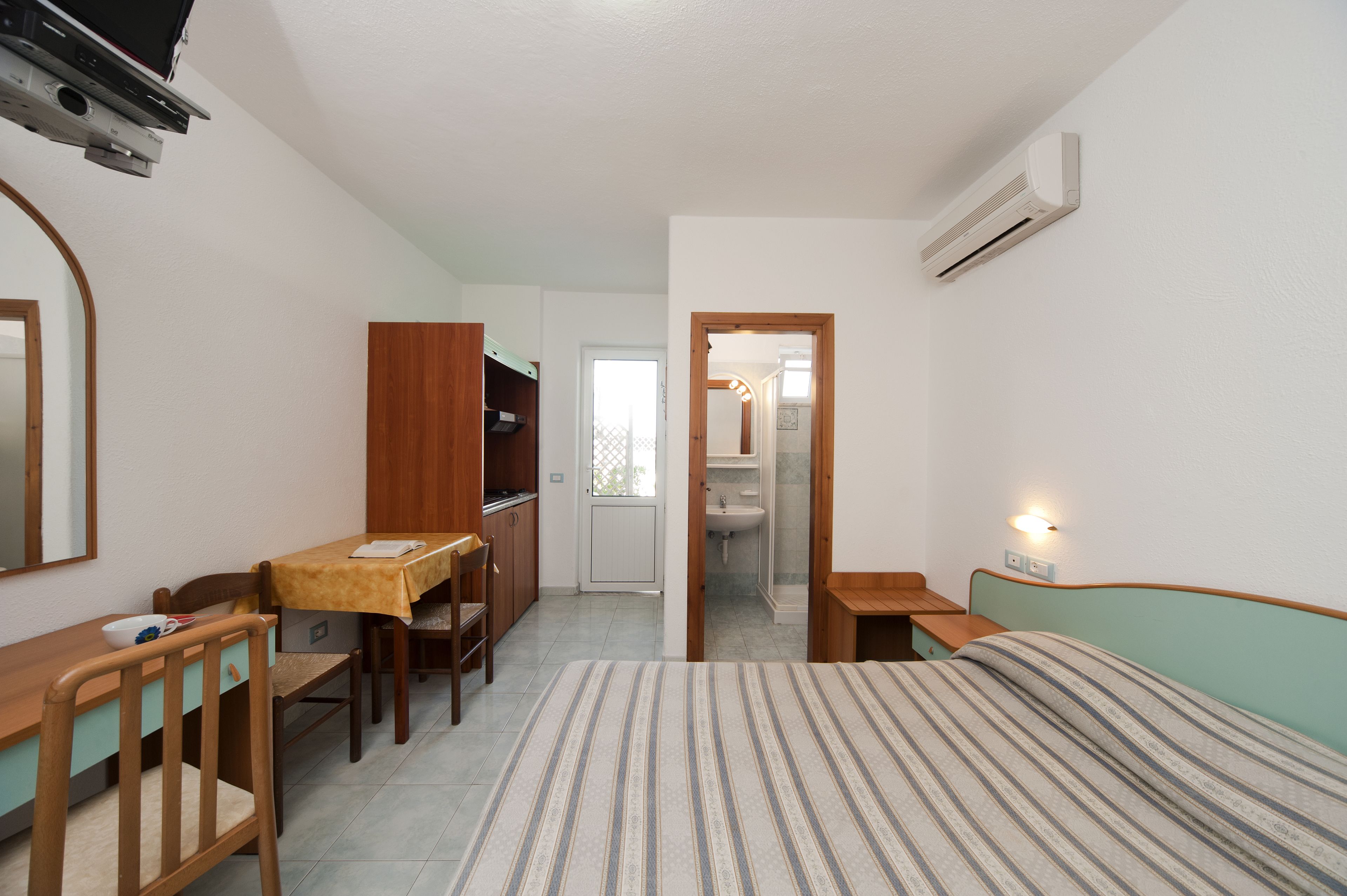 Une petite pièce bien éclairée, avec un lit, un bureau, une chaise, une armoire et une salle de bain visible à travers une porte ouverte.