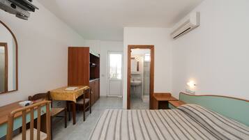 Une petite pièce bien éclairée, avec un lit, un bureau, une chaise, une armoire et une salle de bain visible à travers une porte ouverte.