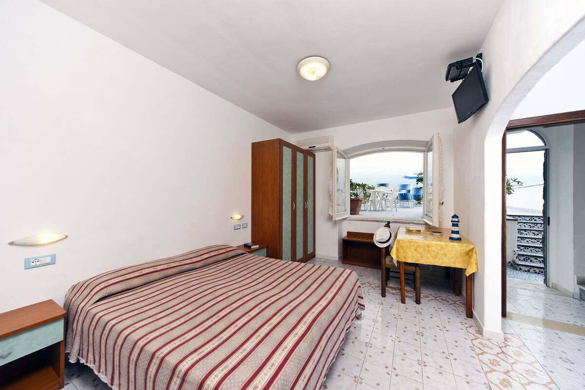 Ein Schlafzimmer mit Bett, Nachttisch, Kleiderschrank, Schreibtisch mit Stuhl und Meerblick durch ein Fenster.