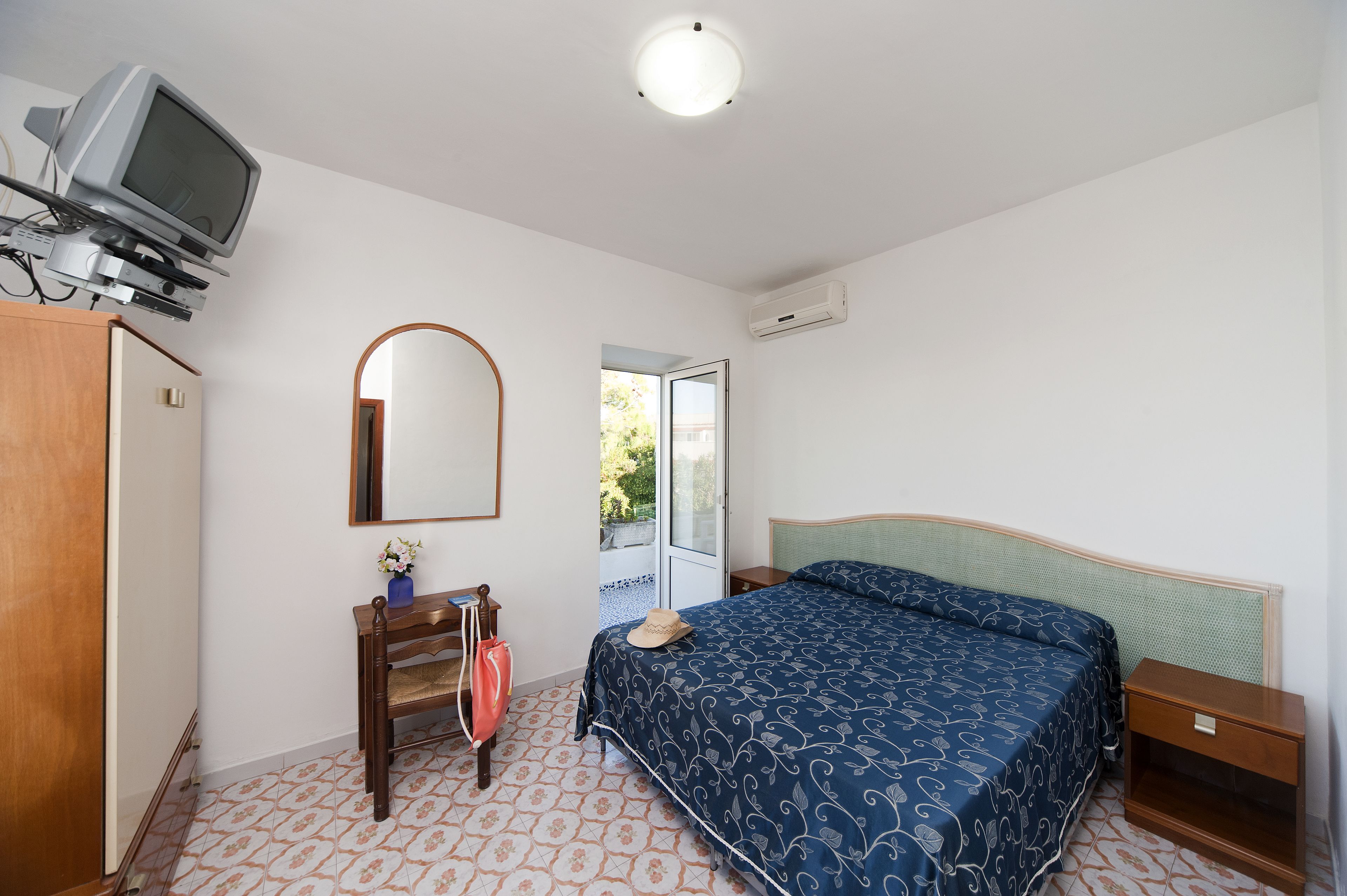 Un dormitorio con cama, televisor montado en la pared, espejo y puerta que da a un balcón.