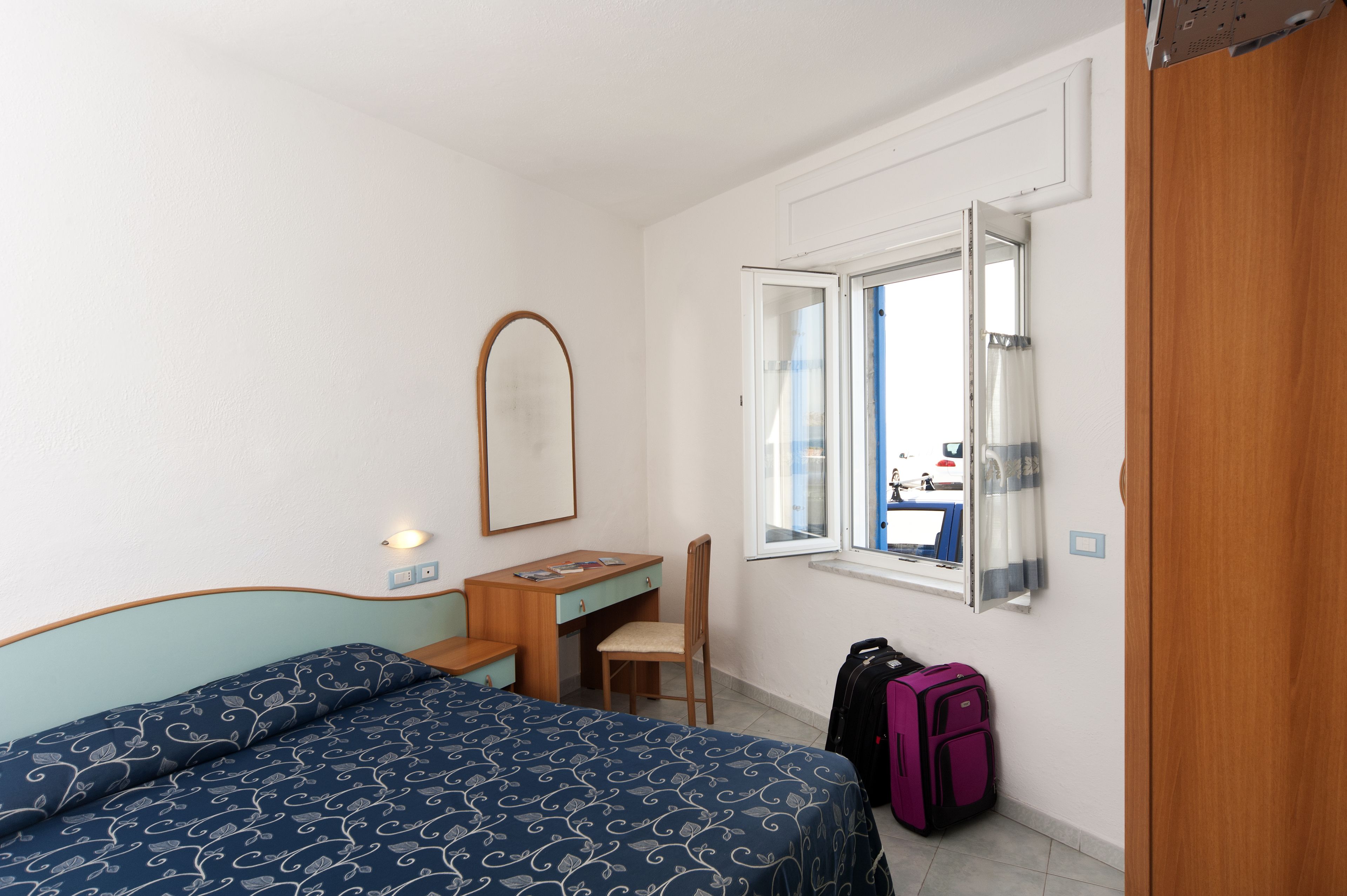 Un dormitorio con cama, escritorio, silla, ventana y maleta.