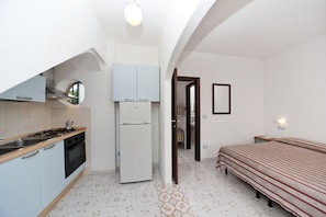 A compact kitchen with a stove, oven, and refrigerator, a bed with a striped bedspread, and a hallway leading to another room. - La Rotonda Sul Mare (Forio)