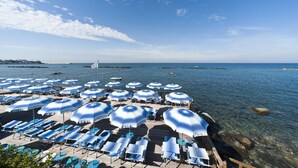 A beachside area with rows of sun loungers and blue umbrellas, overlooking a calm sea.