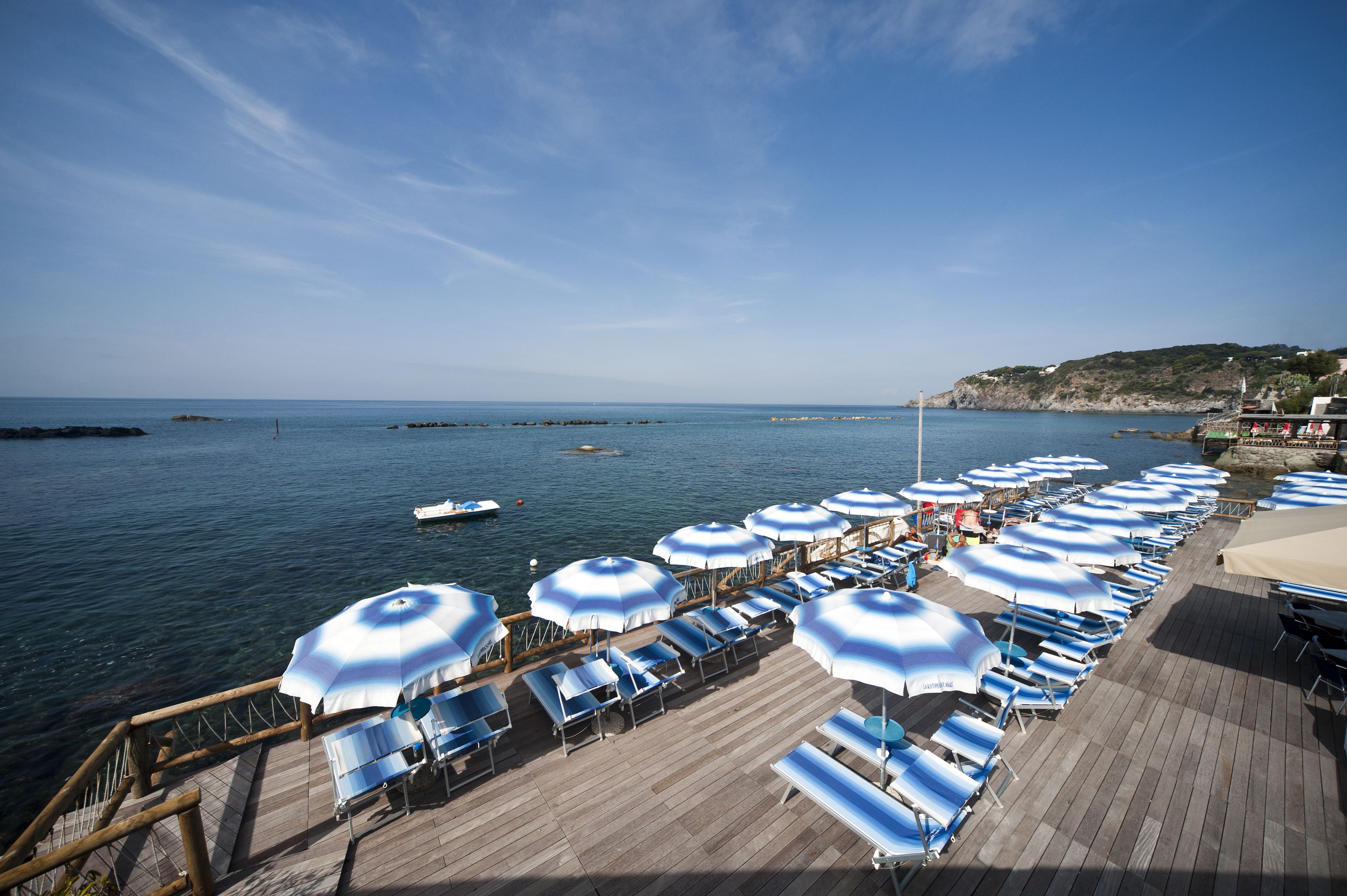 Une terrasse en bois, bordée de rangées de parasols bleus et blancs et de chaises longues, donnant sur une mer calme.