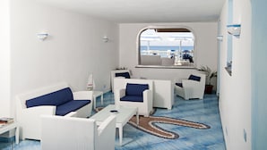 A sunlit room with white wicker furniture, a tiled floor, and a view of the sea through a window.