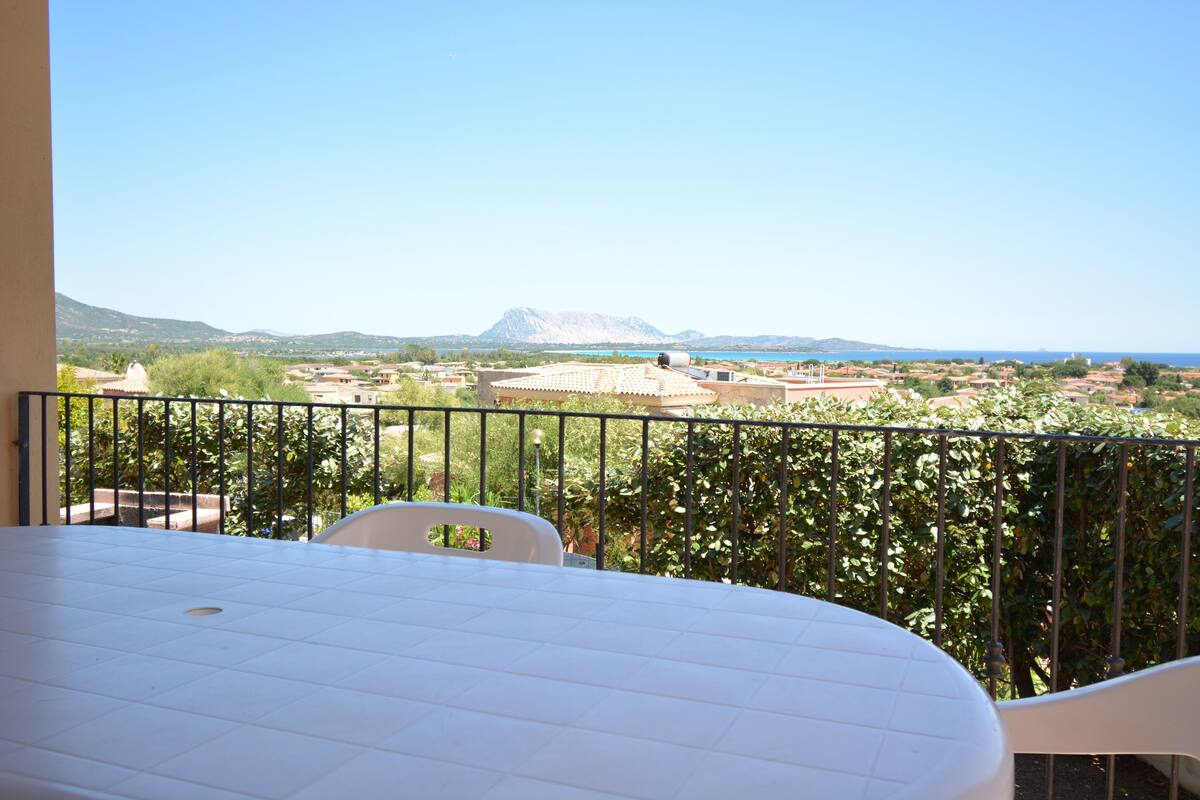 Varanda com mesa e cadeiras, com vista para uma paisagem com edifícios e uma montanha.