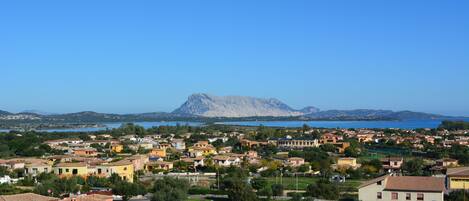 A coastal town with terracotta-roofed buildings, a prominent mountain in the background, and a clear blue sky.