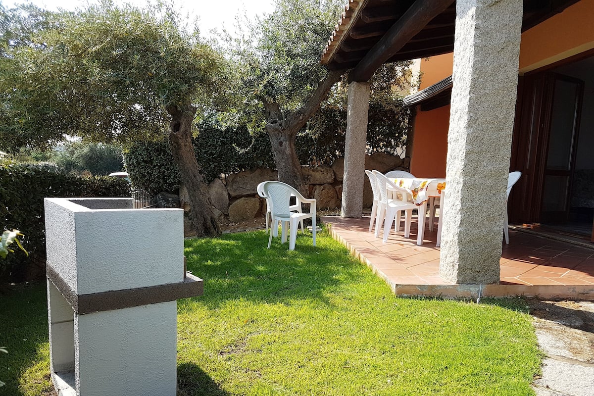 Uma área de pátio com uma mesa e cadeiras de pedra, um vaso de concreto e um muro de pedra ao fundo.