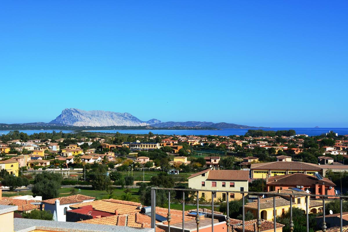 Uma cidade costeira com edifícios de telhados em tons de terracota, uma montanha imponente ao fundo e um céu azul límpido.