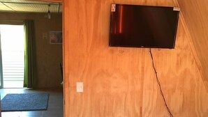 A room with a wooden ceiling, a bed with a red blanket, a television mounted on the wall, and a door leading to another room. - Cabaña en Algarrobo 3 El Canelo (Algarrobo)
