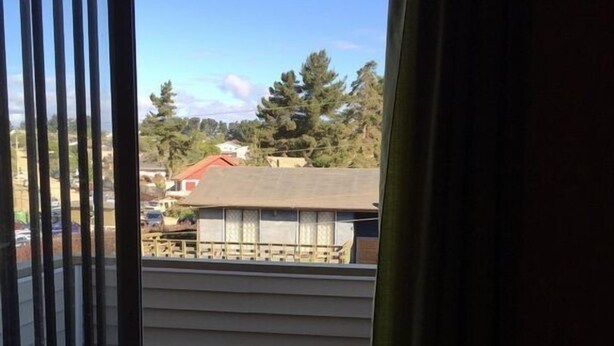 A view through a sliding glass door with a metal railing, leading to a balcony with a tiled floor and a building with a red roof in the background. - Cabaña en Algarrobo 3 El Canelo (Algarrobo)