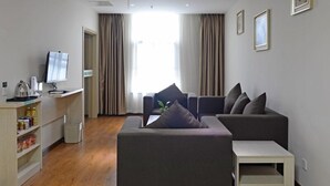 A modern living room with a grey sofa, a white coffee table, and a TV on a stand. There are curtains on the windows, and a small shelf with items on the left. - GreenTree Alliance Suzhou Zhangjiagang Huachang Road Bus Station Hotel (Suzhou)