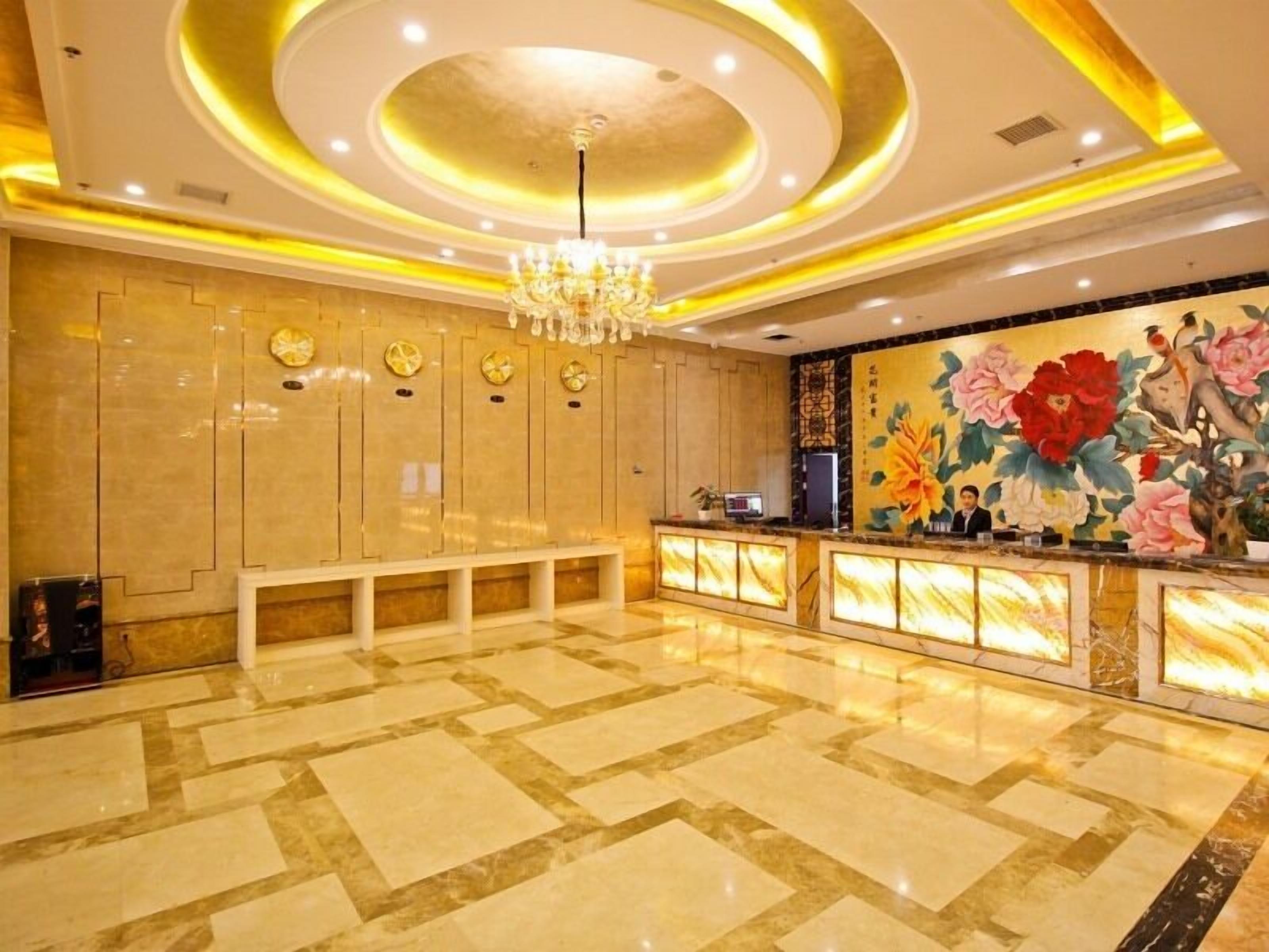 Un vestíbulo de hotel con piso de mármol, una recepción con un mural floral, un candelabro y una fila de relojes en la pared.
