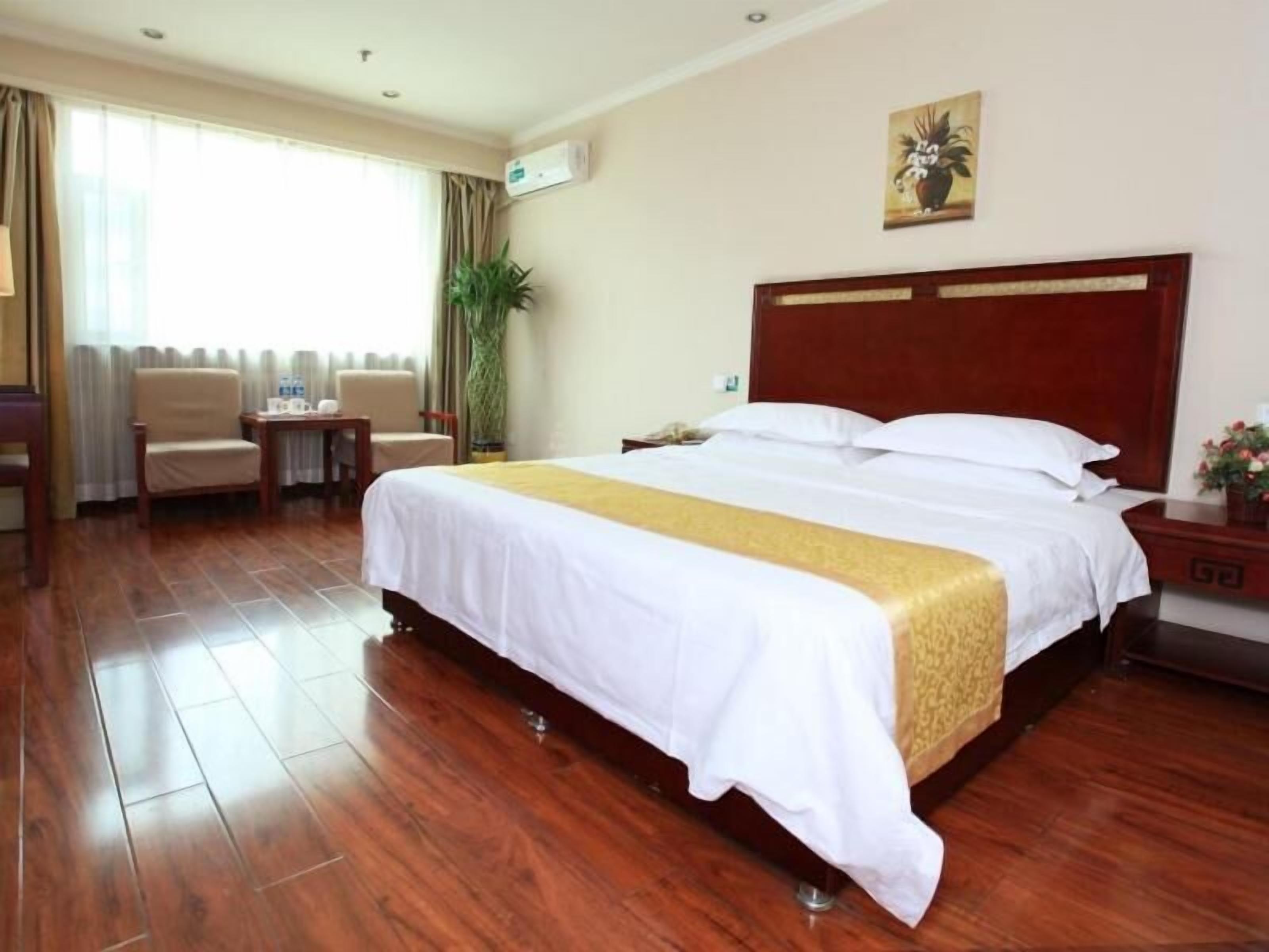 A hotel room with a large bed, a wooden headboard, a bedside table, a chair, a small table, and a potted plant.