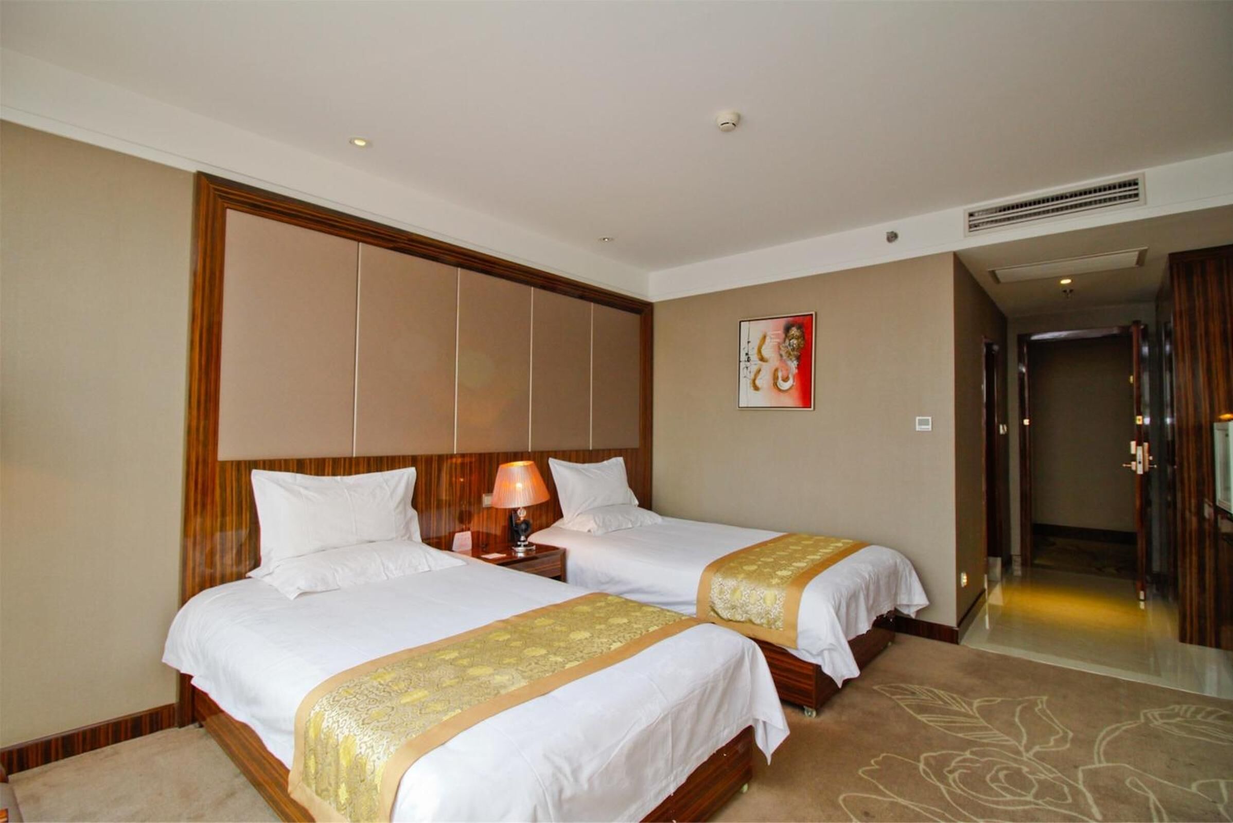 A hotel room with two beds, a wooden headboard, a nightstand with a lamp, a framed picture on the wall, and a carpet with a floral pattern.