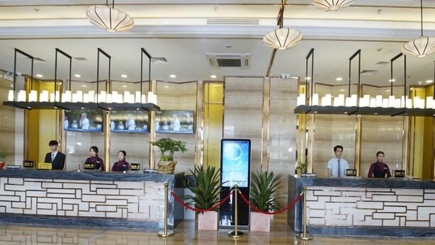 A hotel reception area with two reception desks, staff members, and a digital information board.