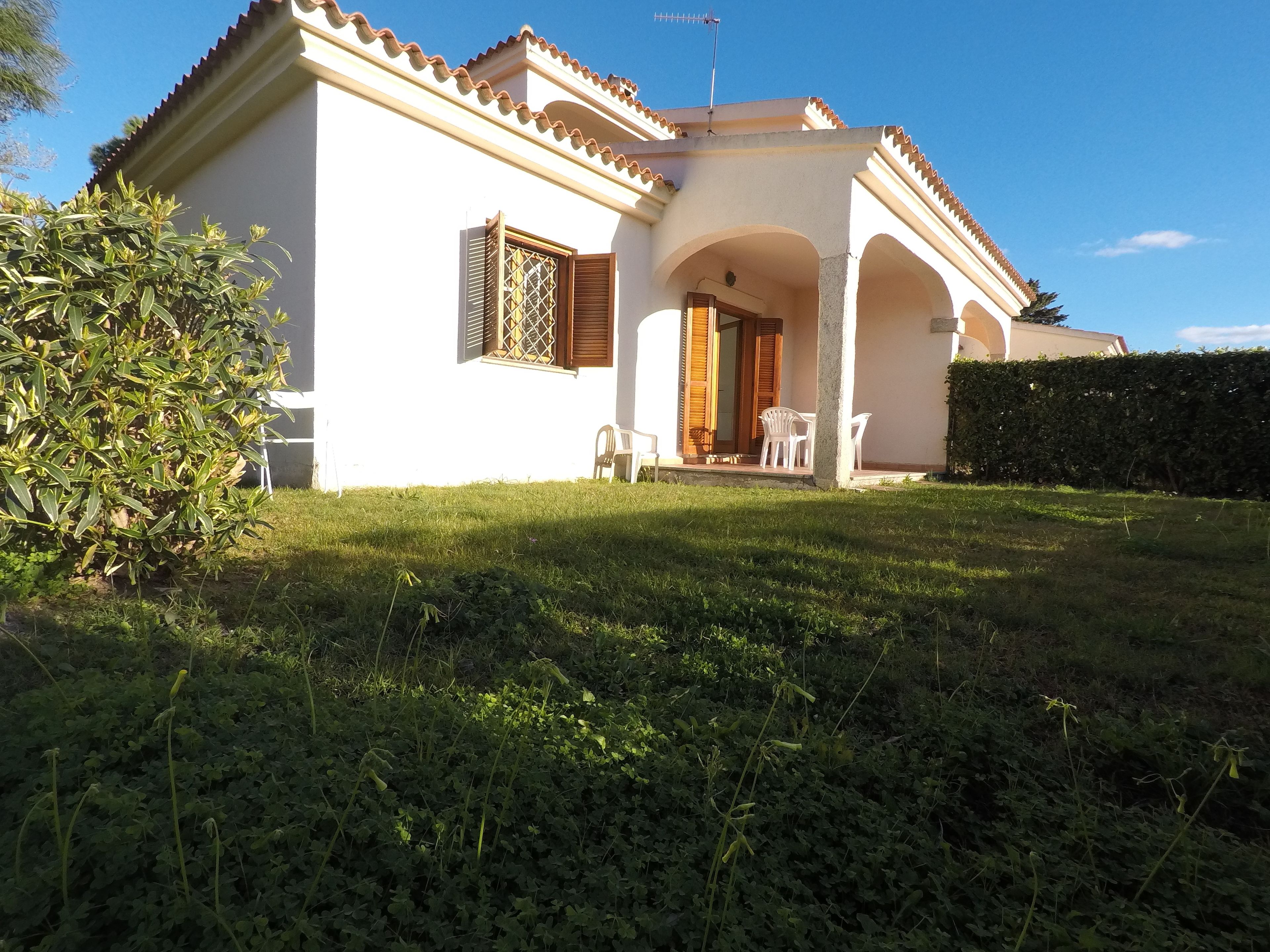 A white villa with a tiled roof, a covered patio with chairs, and a well-maintained lawn.