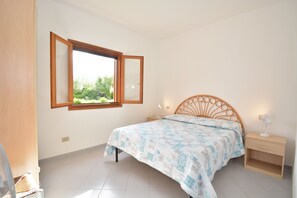 Un dormitorio con cama, mesita de noche, ventana con contraventana abierta y vista a zonas verdes.