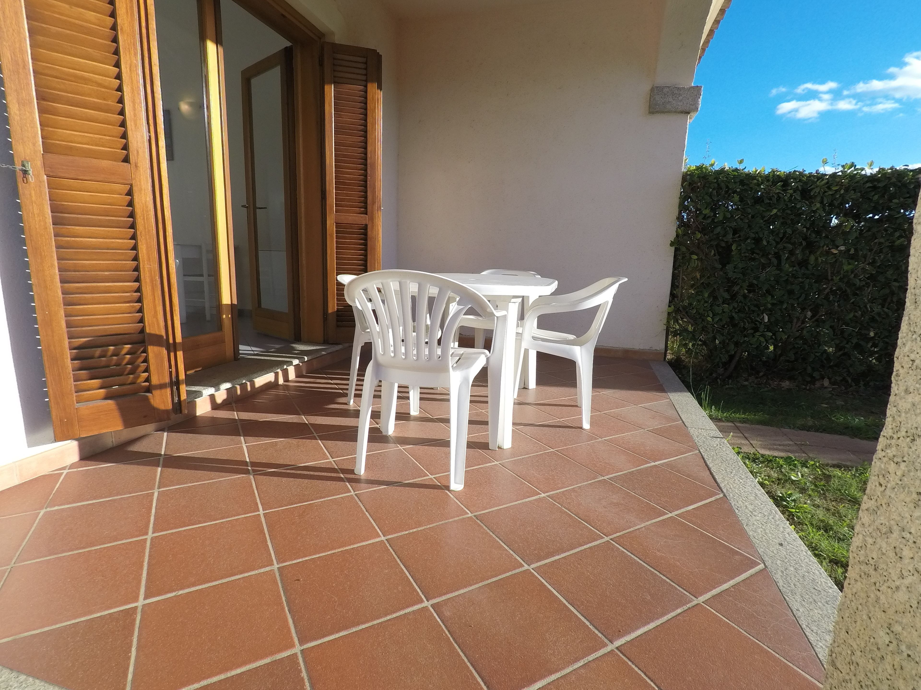 Une terrasse avec un sol carrelé, des chaises en plastique blanches et une porte en bois dotée de volets.