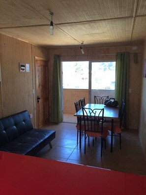 A room with a wooden ceiling, a dining table with chairs, a black sofa, and a window with green curtains. - Cabaña en Algarrobo 4 El Canelo (Algarrobo)