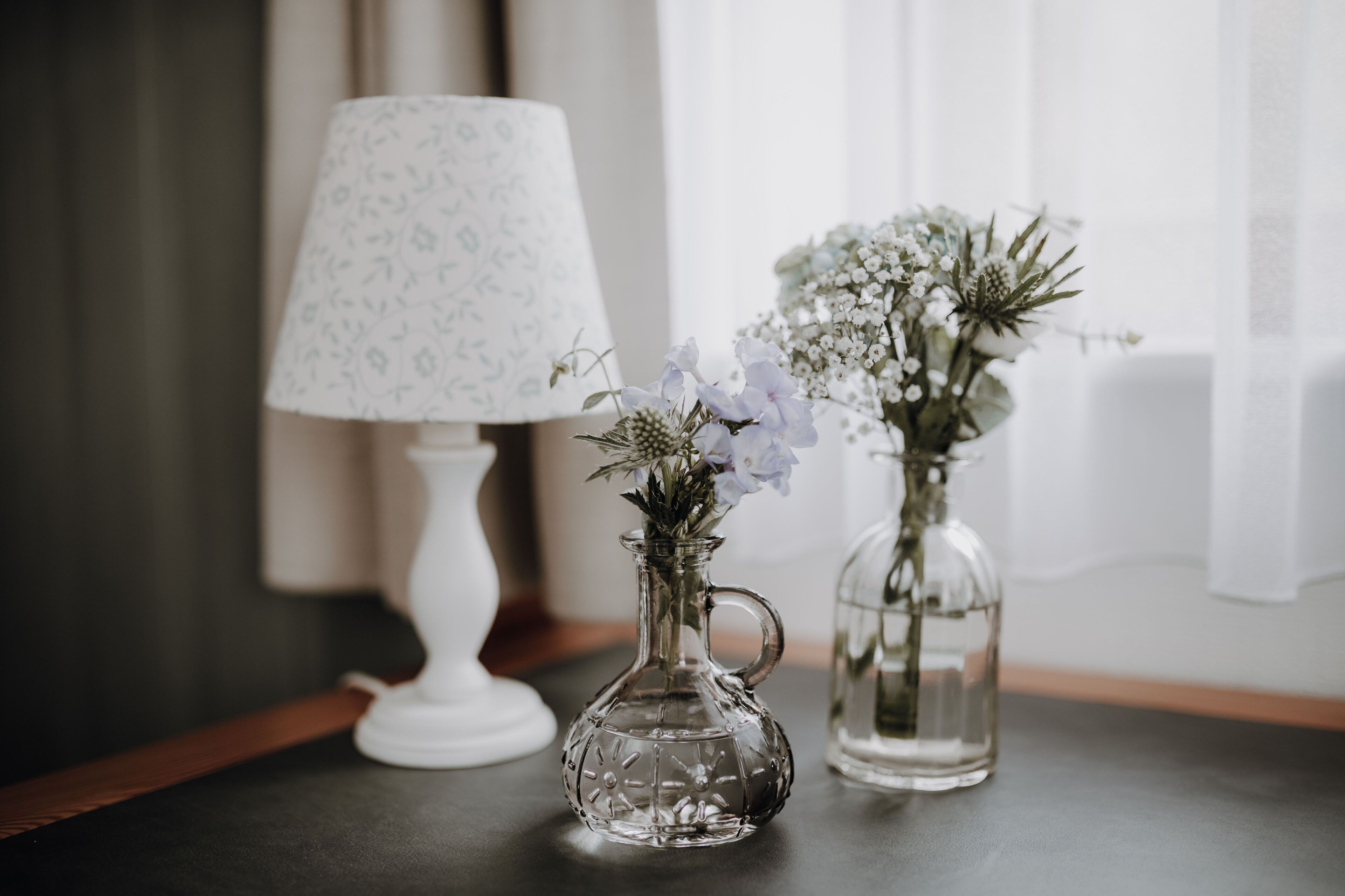 Two glass vases with flowers on a table, a patterned lamp, and a window with sheer curtains.