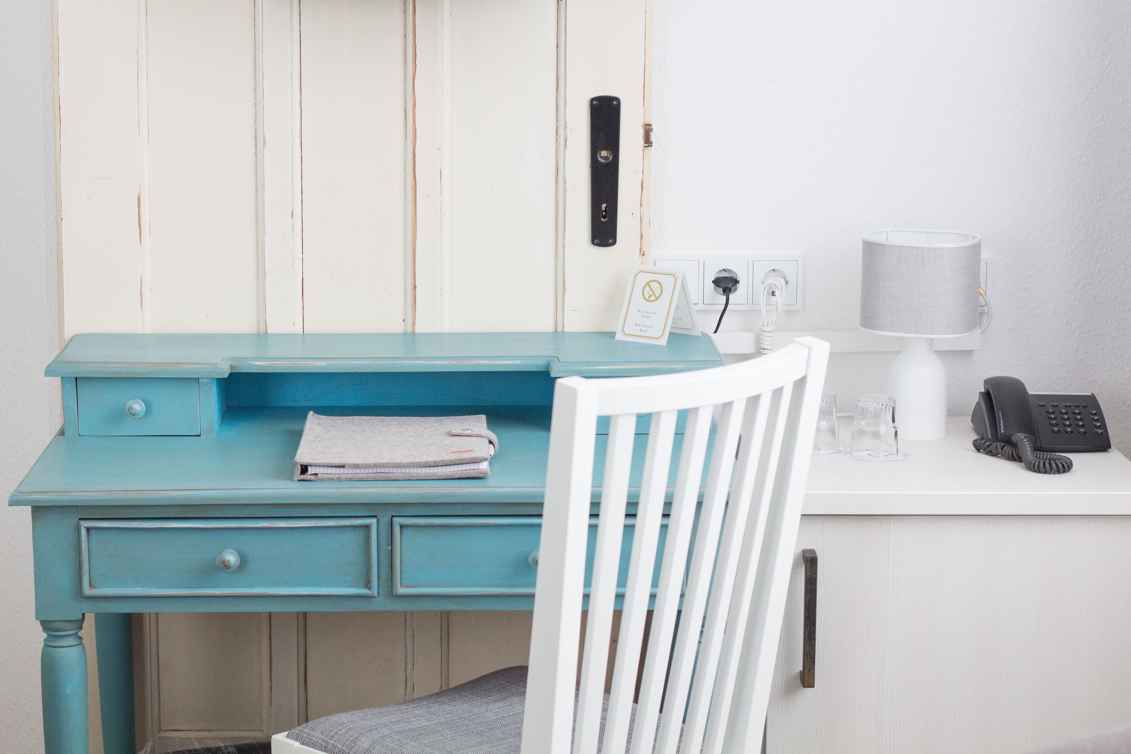 A teal desk with a white chair, a telephone, a lamp, and a sign on the wall.
