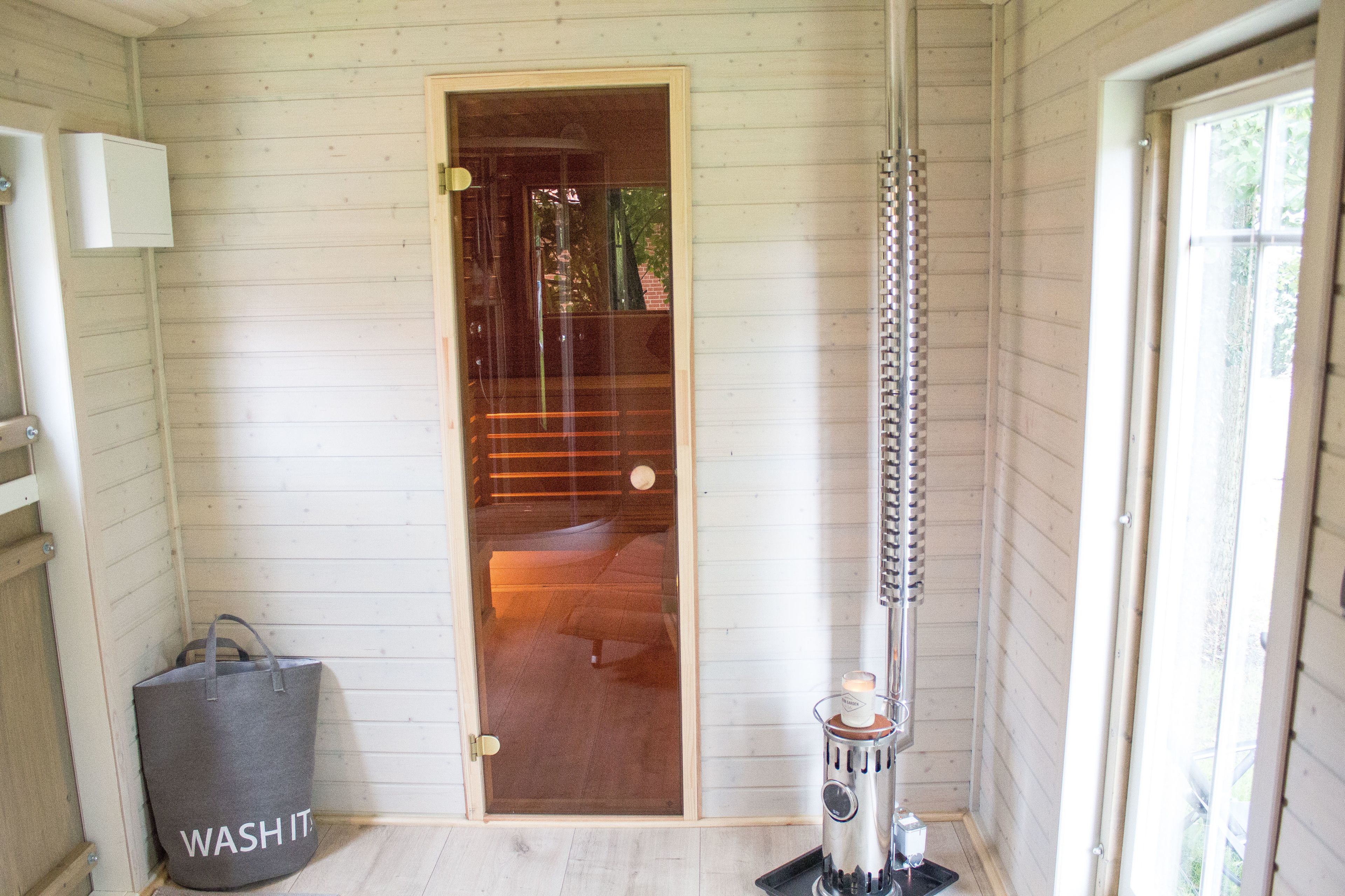 A room with wooden walls, a door, a grey bag labeled "WASH IT", and a tall, cylindrical heater.