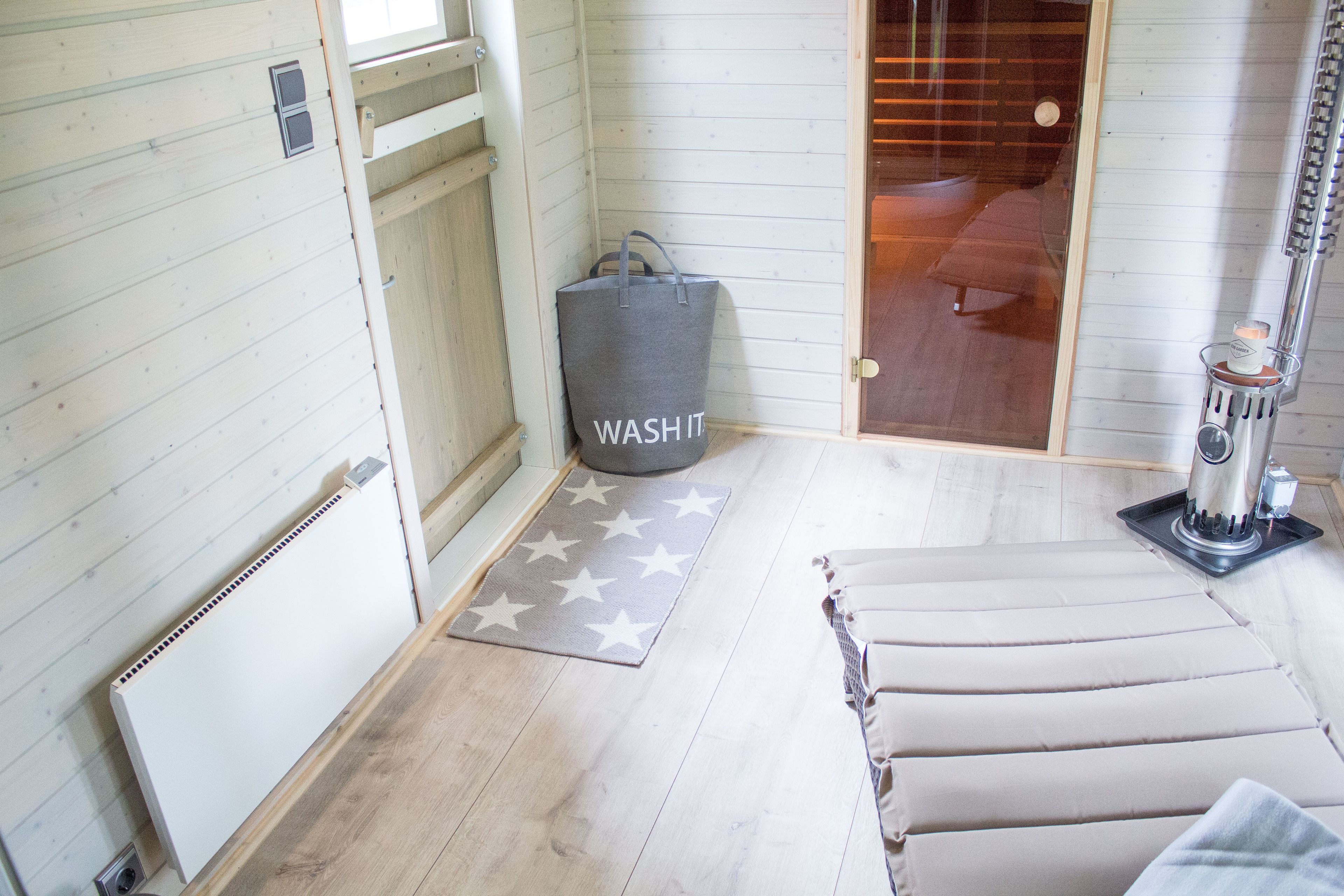 A bathroom with a wooden floor, a towel rack, a radiator, a star-patterned rug, a grey bag, and a shower area.
