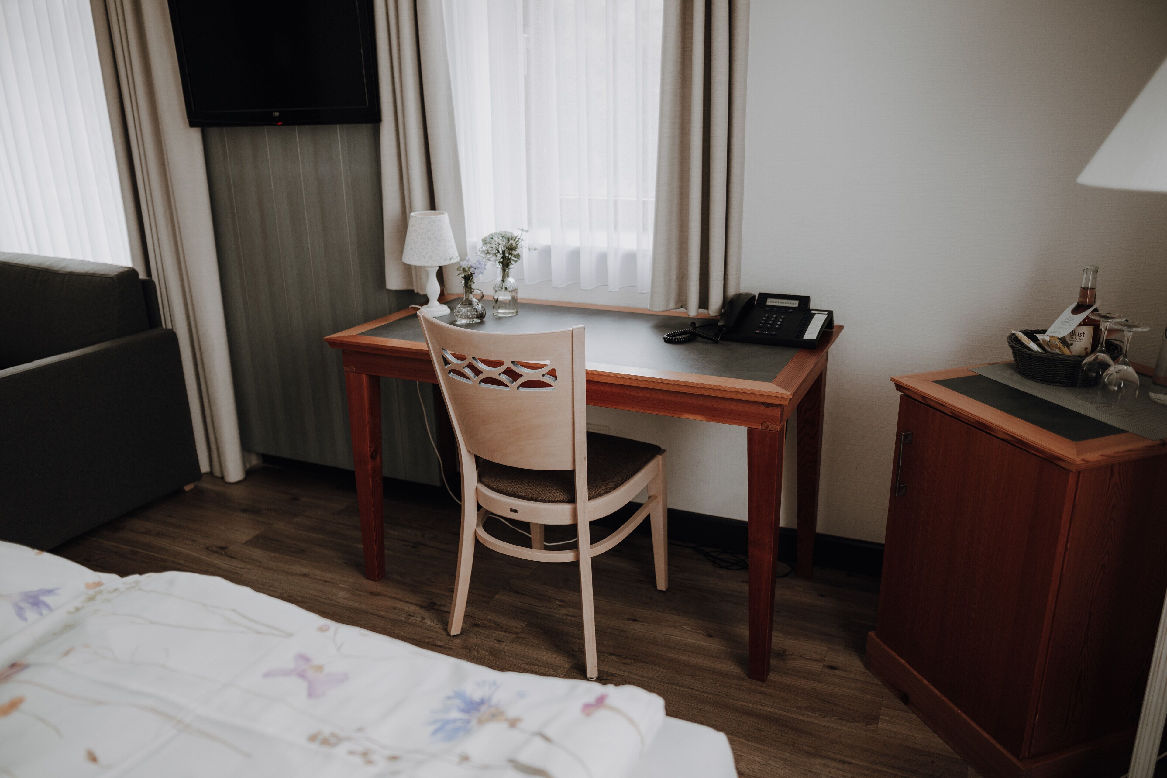 A hotel room with a wooden desk, a chair, a bed with a floral bedspread, a television, and a minibar.