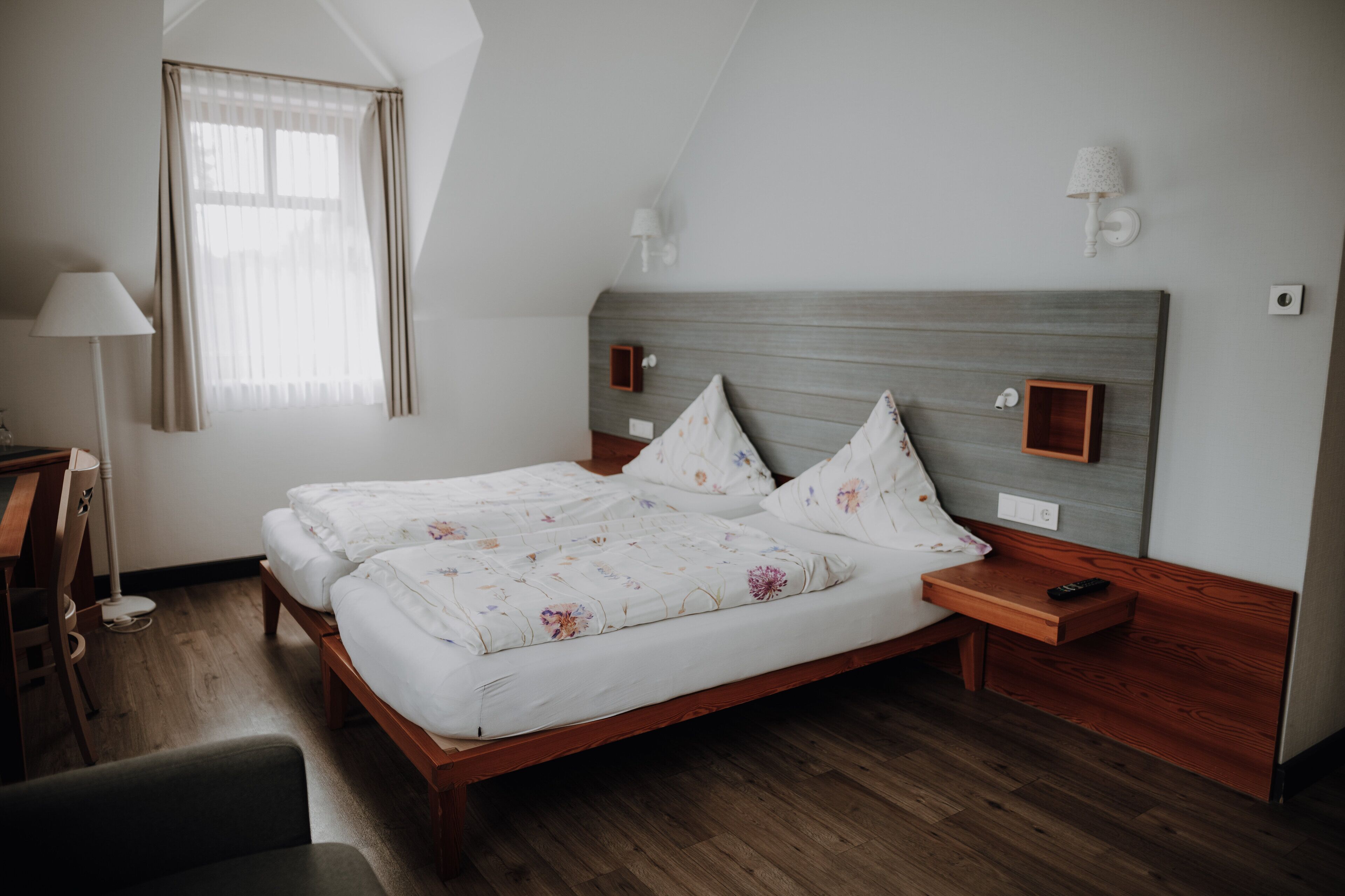 Ein Doppelbett mit Holz Kopfteil, zwei Nachttische, eine Lampe und ein Fenster mit Vorhängen.