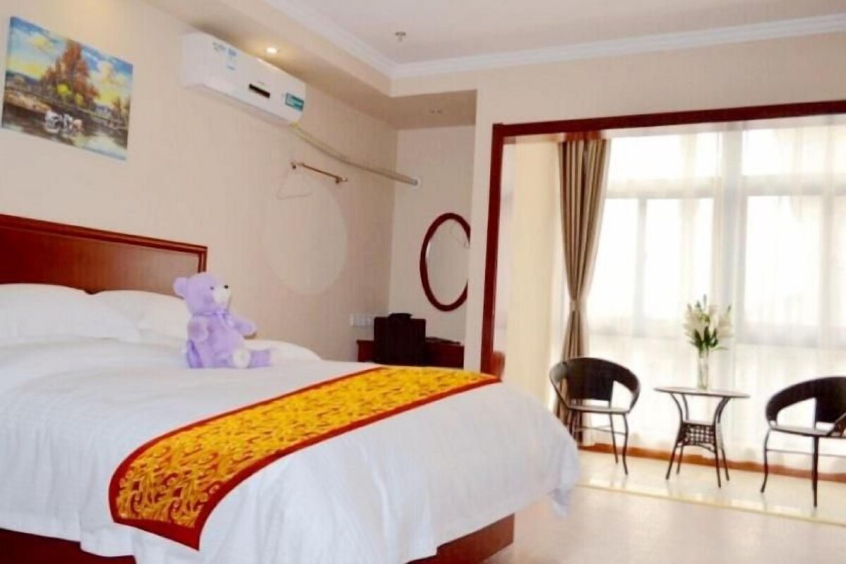 Quarto de hotel com cama, um urso de pelúcia, um quadro, ar condicionado, uma escrivaninha e duas cadeiras.