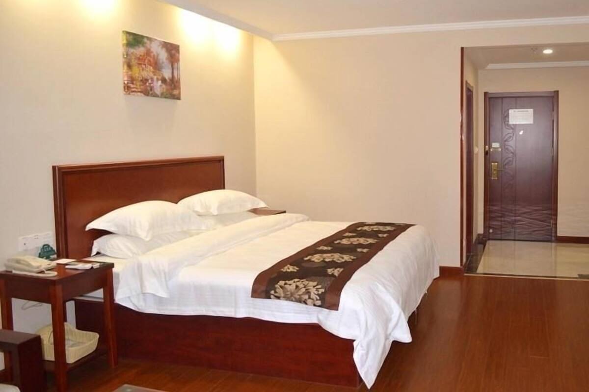 Une chambre d’hôtel avec un lit en bois, du linge de lit blanc, une table de chevet, un téléphone et un tableau au mur.