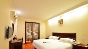 Quarto de hotel com piso de madeira, uma cama com roupa de cama branca, escrivaninha com cadeira, televisão presa na parede e janela com cortinas.
