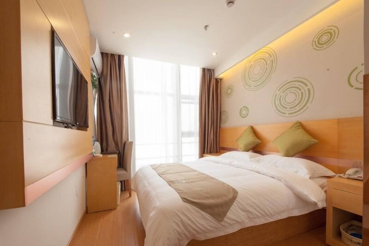 Ein modernes Hotelzimmer mit einem großen Bett, einem Nachttisch, einem an der Wand montierten Fernseher und einem Fenster mit Vorhängen.