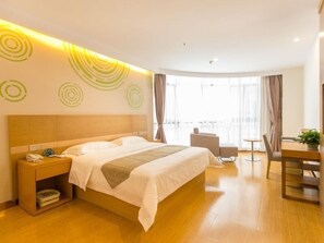 A hotel room with a large bed, a bedside table with a phone and a basket, a small round table with a chair, and a view of the city through large windows.