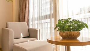 A modern living room with a beige armchair, a round wooden table with a potted plant, and sheer curtains allowing natural light. - Greentree Inn (Suzhou)