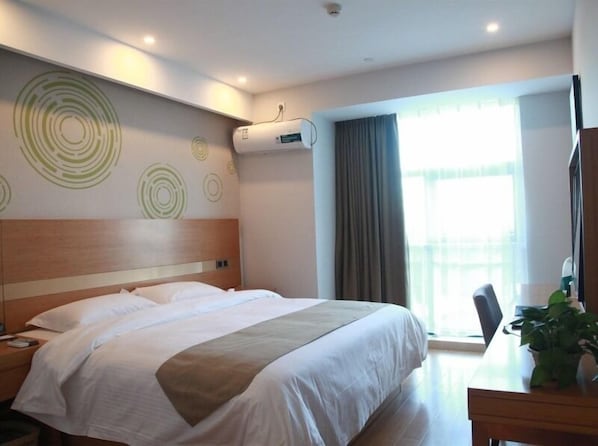 A hotel room with a large bed, a desk with a chair, a potted plant, and a window with curtains.