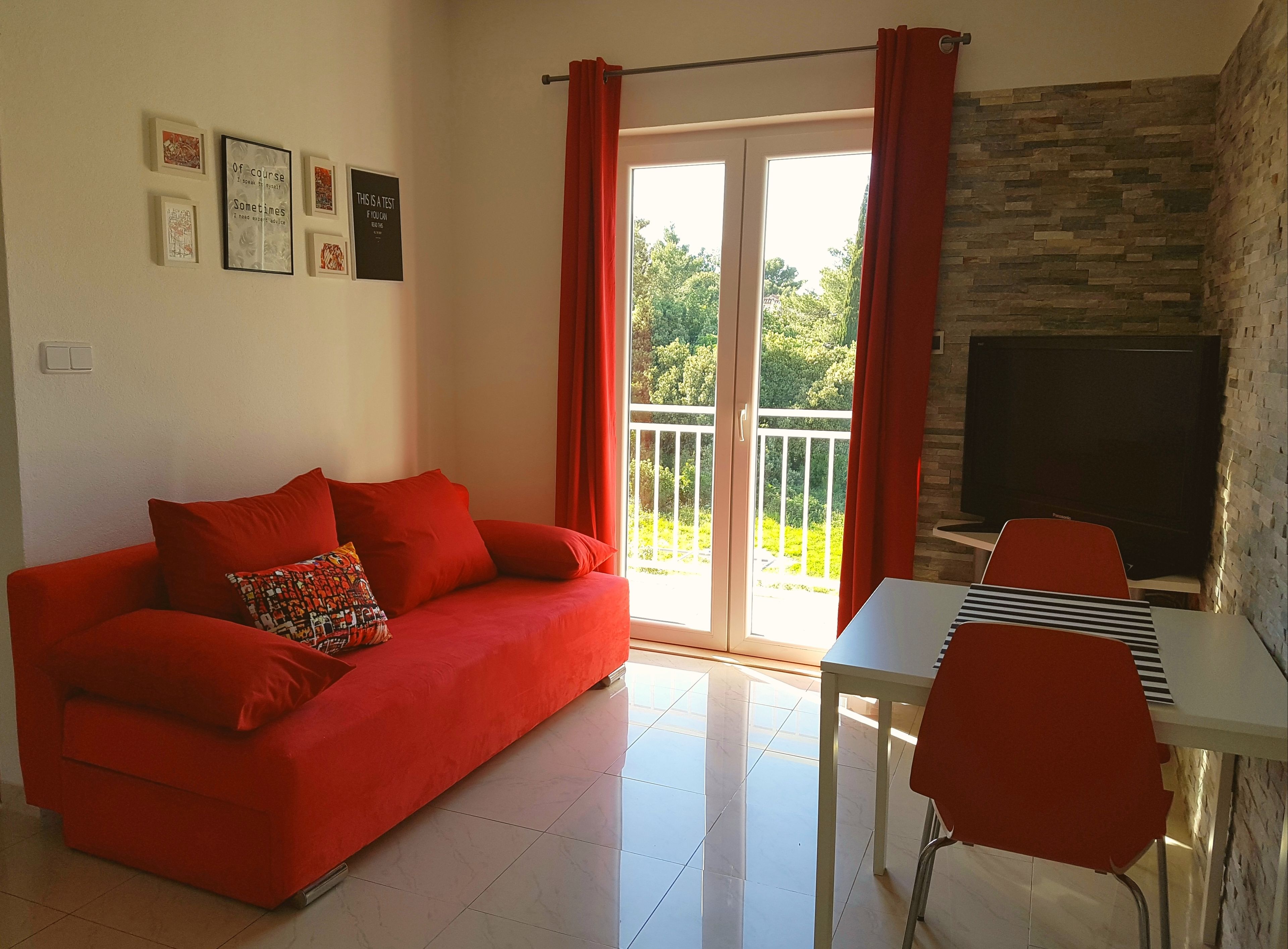 A living room with a red sofa, a white table with a red chair, a large TV, and a balcony with red curtains.