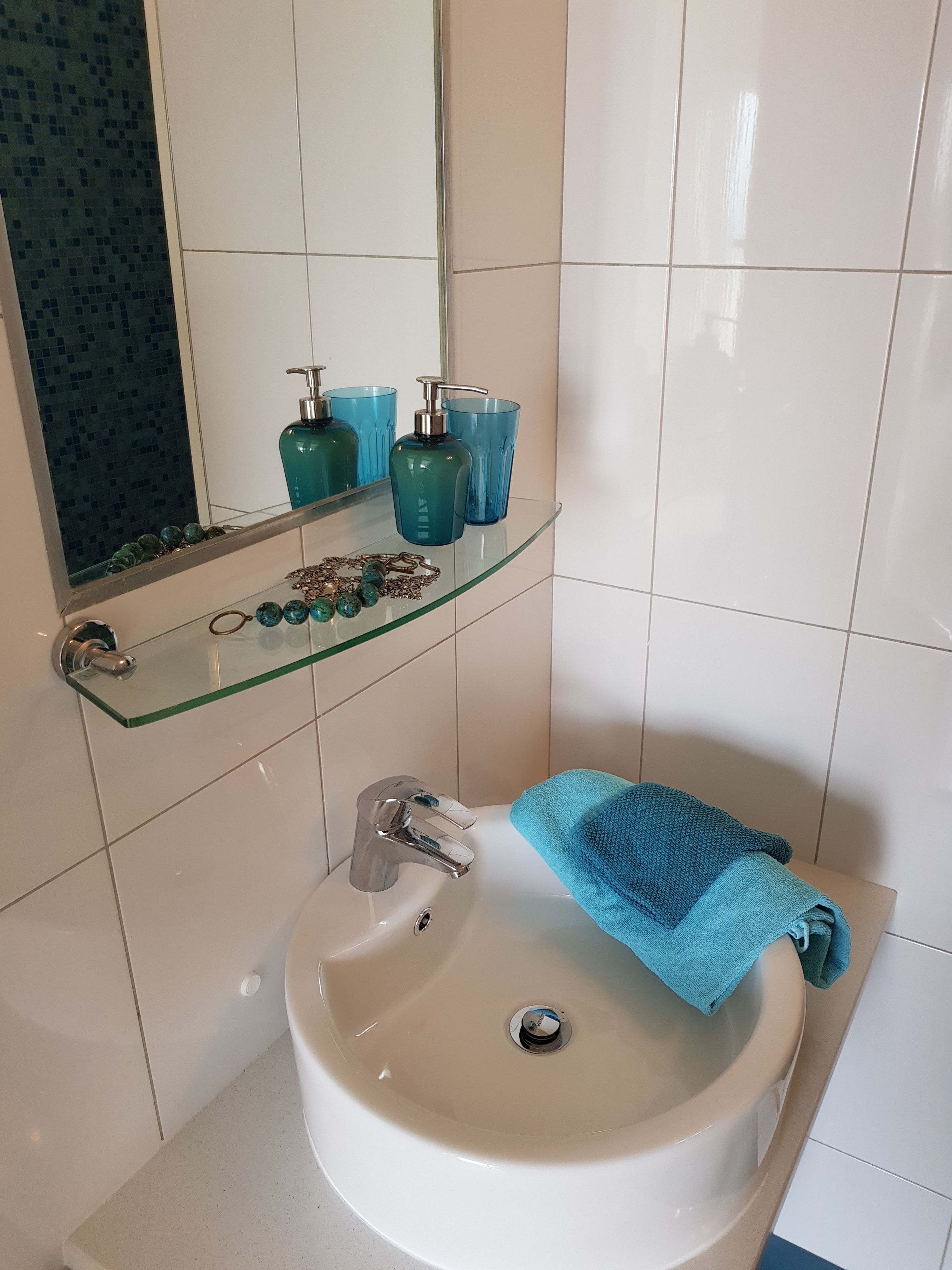 Un baño con lavamanos blanco, estante de vidrio con artículos de aseo y toalla azul.
