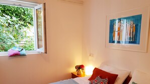 Un dormitorio con cama, almohadas, una mesita de noche con lámpara, una ventana con vistas a la vegetación y un cuadro en la pared.