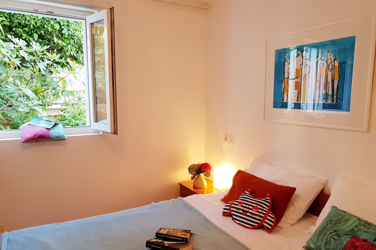Een slaapkamer met een bed, kussens, een nachtkastje met een lamp, een raam met uitzicht op groen en een schilderij aan de muur.