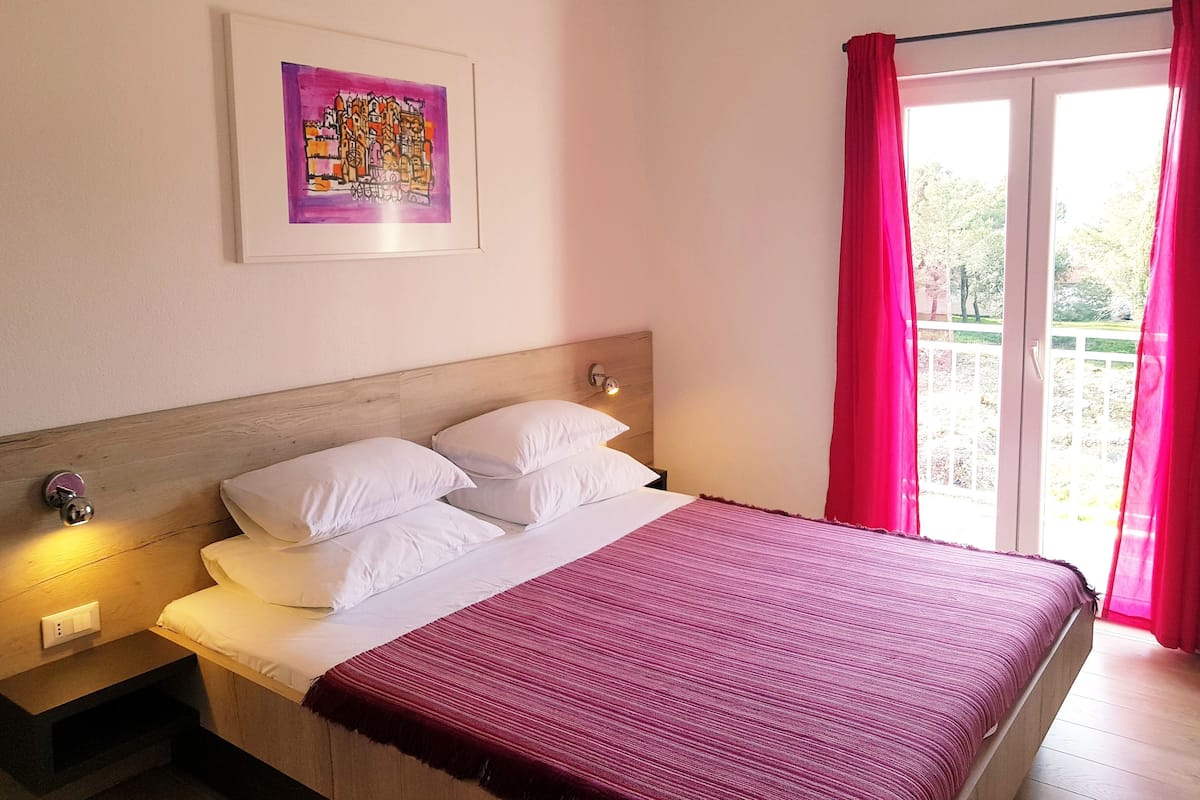 Een slaapkamer met een bed, witte kussens, een houten hoofdbord, een ingelijst schilderij aan de muur en een raam met gordijnen.