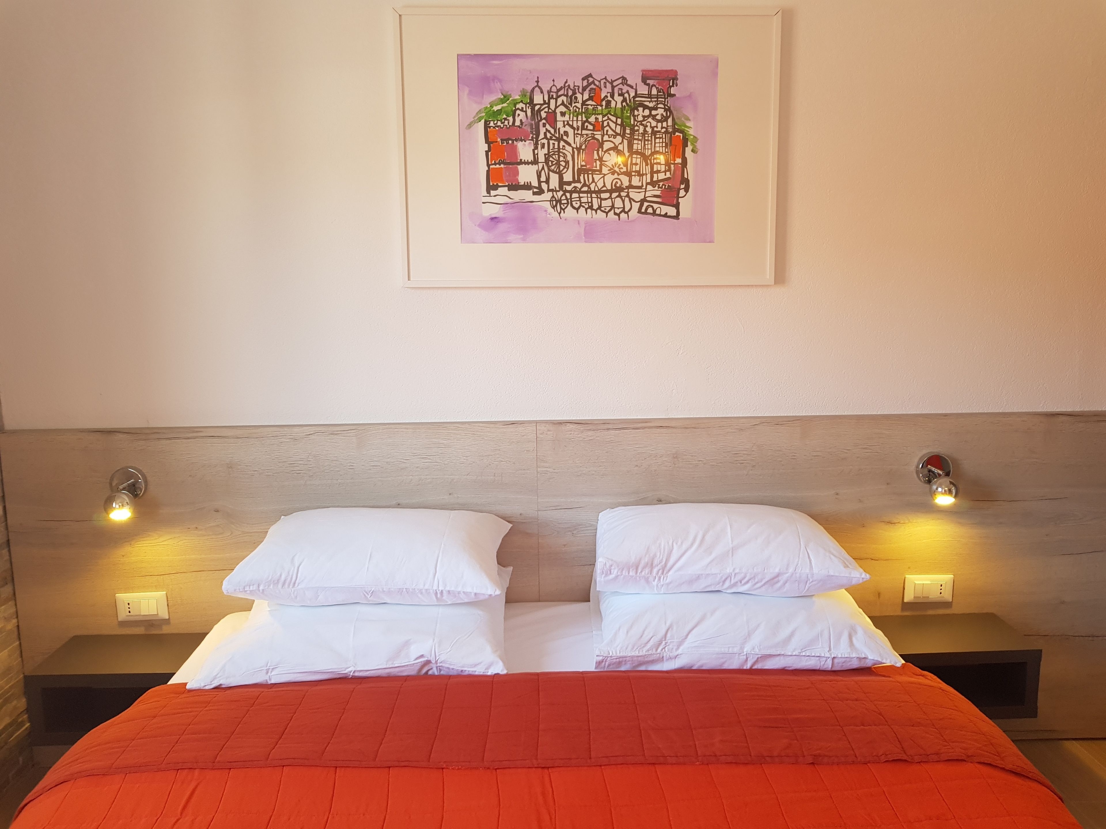 A bedroom with a wooden headboard, two bedside tables with wall-mounted lamps, a red bedspread, and a framed artwork on the wall.