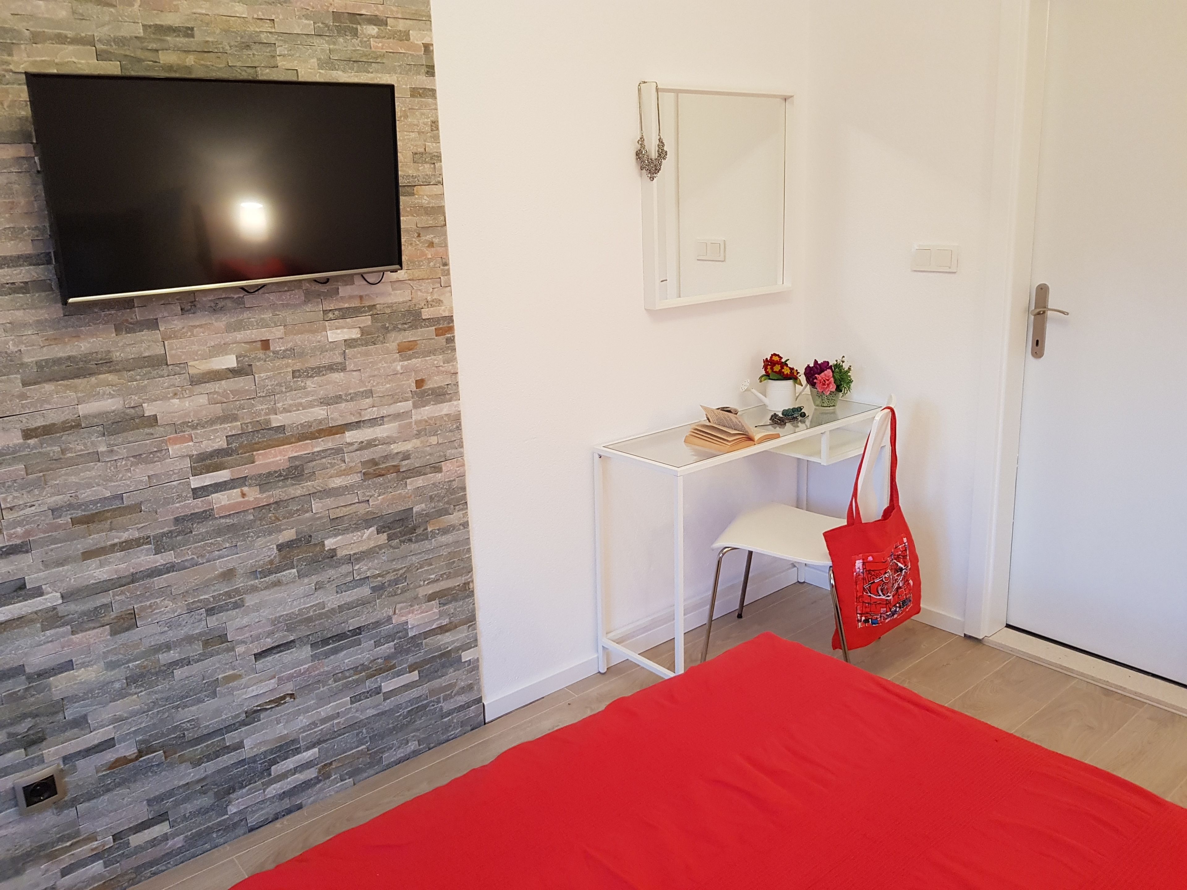 A room with a stone wall, a flat-screen TV mounted above a desk, a mirror, a door, and a red bedspread.