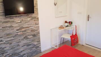 A room with a stone wall, a flat-screen TV mounted above a desk, a mirror, a door, and a red bedspread.
