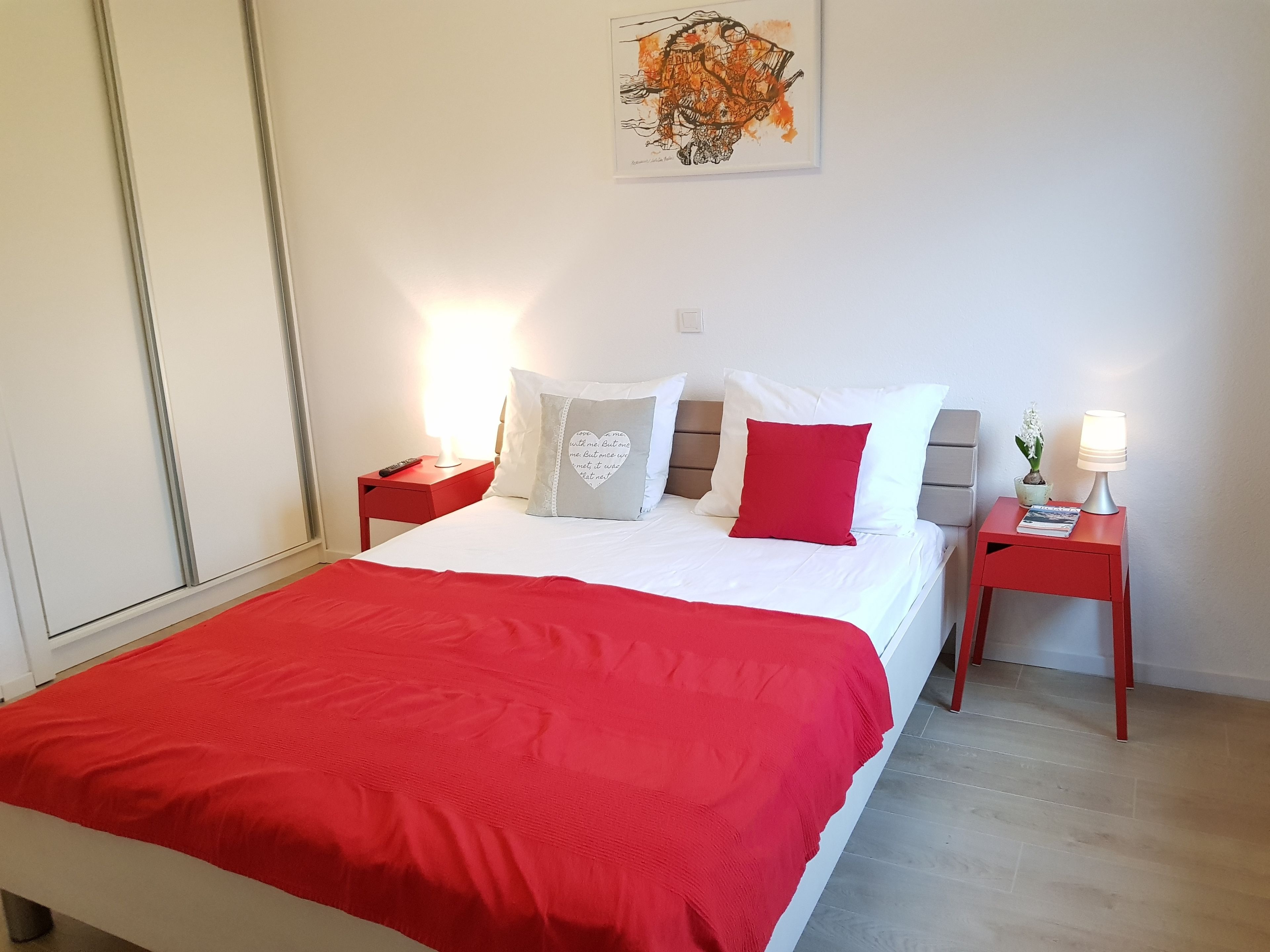 Una cama hecha con cuidado, con un edredón rojo, almohadas blancas y una almohada roja con un diseño de corazón. Hay dos mesitas de noche, una con una lámpara y la otra con un control remoto.