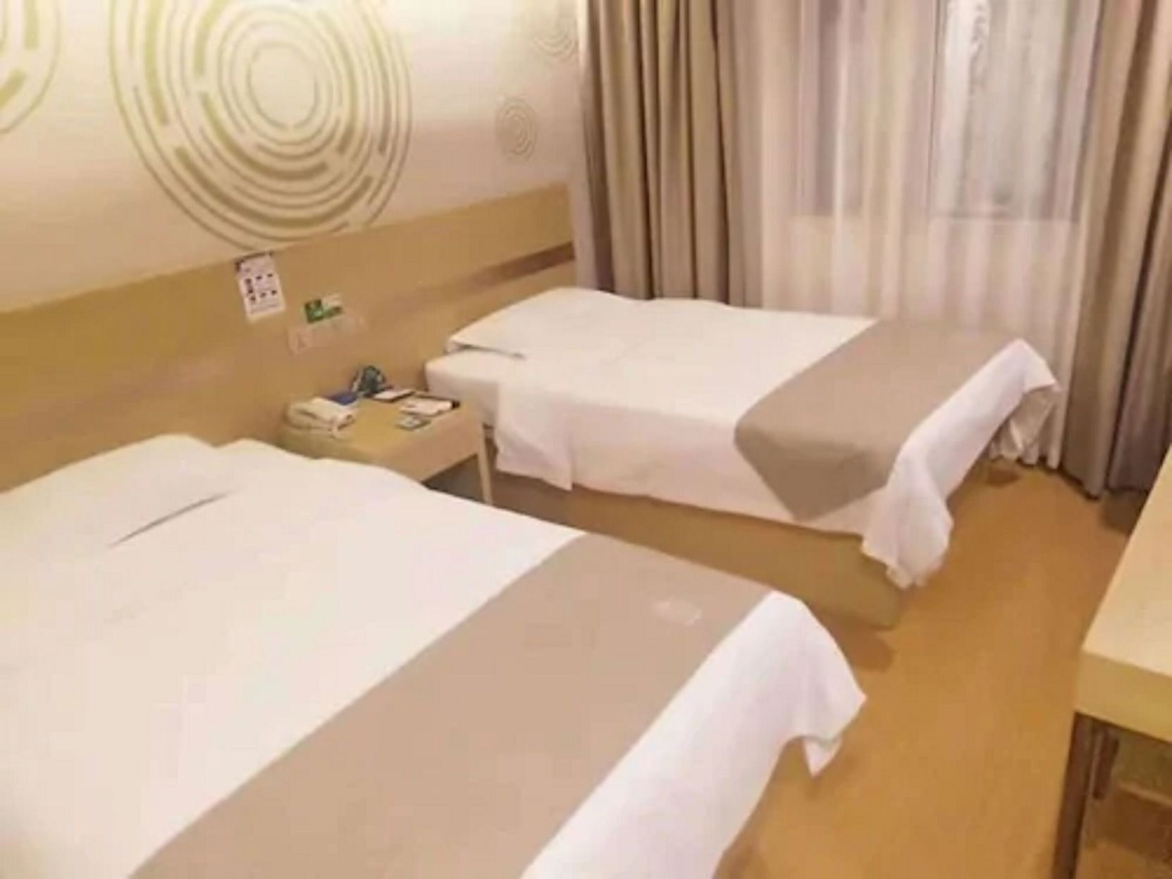 A hotel room with two beds, a bedside table with a phone and a book, and a curtain.