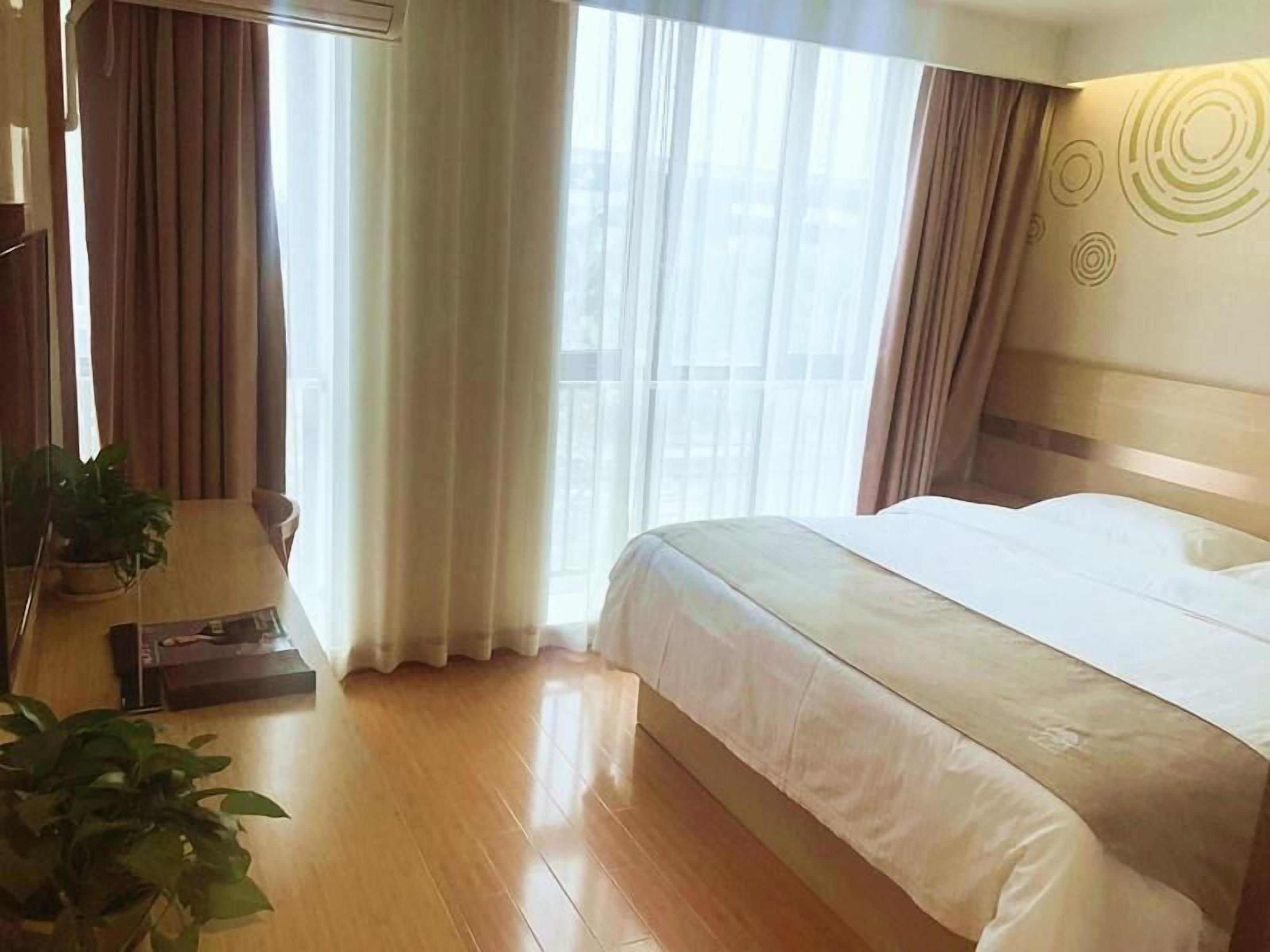 A hotel room with a large bed, a wooden floor, a small table with a plant, and a window with curtains.