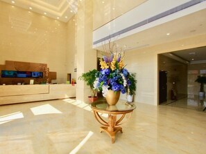 Una hall d'albergo moderna con una reception, un'ampia composizione floreale su un tavolo in vetro e un display digitale a parete.