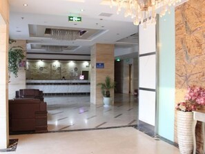 A hotel lobby with a reception desk, a chandelier, a potted plant, and a marble floor.
